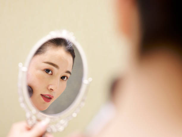 istock beautiful young asian woman looking into mirror while applying make-uo 980583222
