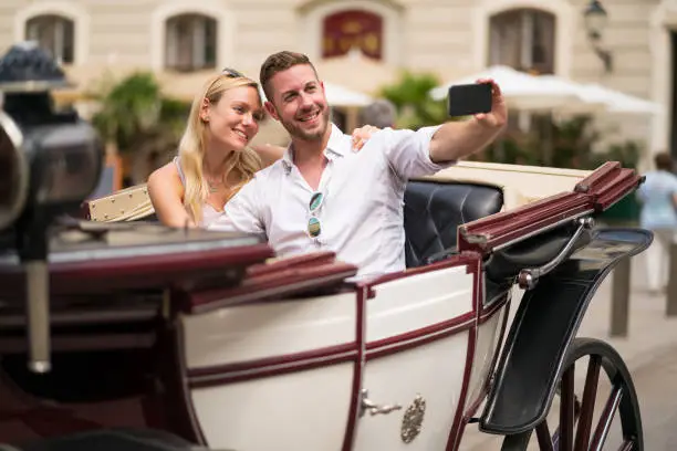 happy young tourist couple sitting in horse carriage on sightseeing tour through Vienna in summer taking selfie souvenir pictures