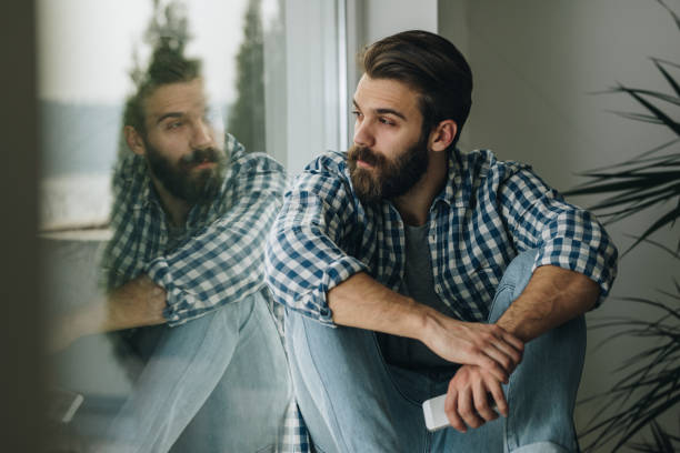 Thoughtful man relaxing on window sill at home. Young pensive man sitting by the window sill and looking through it. soul searching stock pictures, royalty-free photos & images