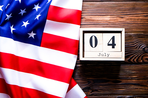 Ruffled American flag and wooden cube calendar with 4th of July, USA Independence Day date on brown wood vintage table, copy space background. US patriotic festive composition, top view, close up.