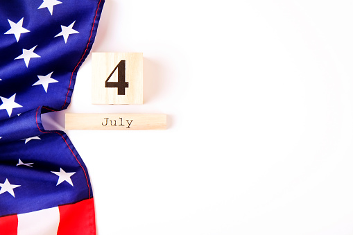 Ruffled American flag and wooden cube calendar with 4th of July date. Happy Independence Day greeting card template on white copy space background. US patriotic festive composition, top view, close up