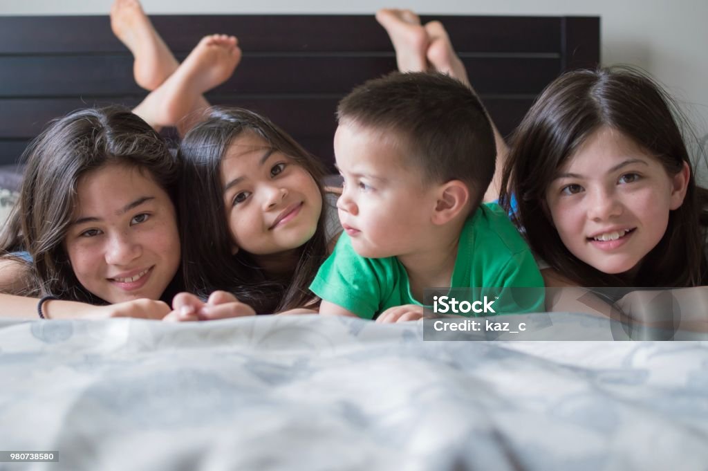 Siblings lying on a bed together A photograph of four Eurasian siblings lying on a bed side by side, on their front with feet in the air. New Zealand Stock Photo