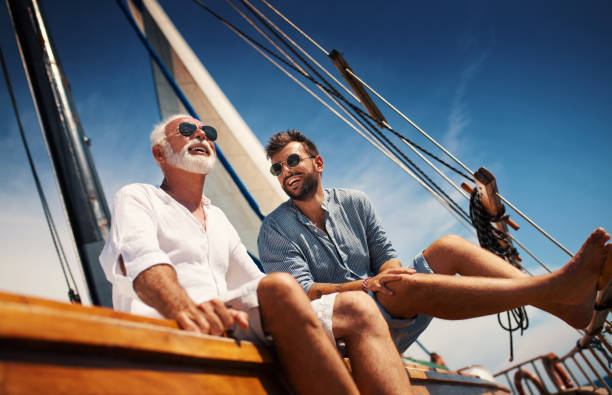 Father and son sailing. Closeup low angle view of mid 60's man and his son enjoying summer holiday on a sailboat. They are sitting on the deck, looking at the distance, talking and laughing. old rich white guy stock pictures, royalty-free photos & images