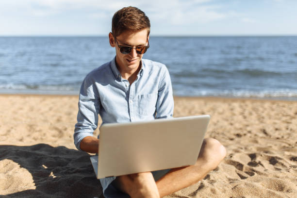 istock Young guy with glasses, working on his laptop on the beach, work on vacation, suitable for advertising, text insertion 980795832