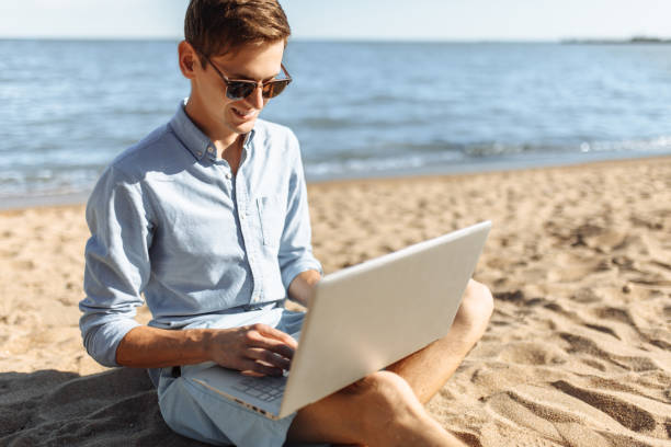 istock Young guy with glasses, working on his laptop on the beach, work on vacation, suitable for advertising, text insertion 980795854