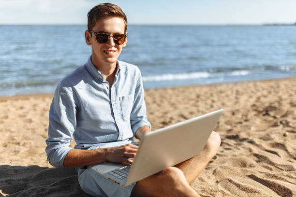 istock Young guy with glasses, working on his laptop on the beach, work on vacation, suitable for advertising, text insertion 980795866