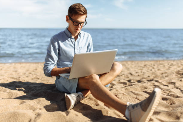 istock Young guy with glasses, working on his laptop on the beach, work on vacation, suitable for advertising, text insertion 980795924