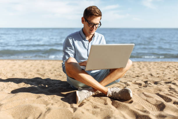 istock Young guy with glasses, working on his laptop on the beach, work on vacation, suitable for advertising, text insertion 980795936