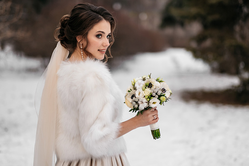 Portrait of a beautiful bride in a winter forest. Woman in a wedding dress, in a white fur coat and with a bouquet in hands