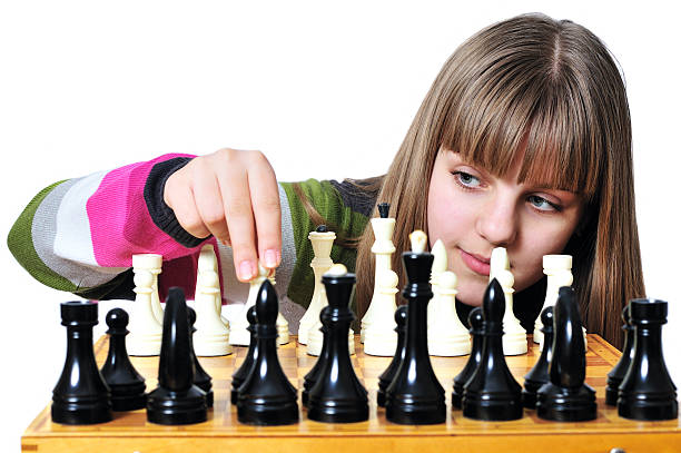 Horizontal photograph of one hand holding and moving a strategic move black queen on a chessboard in game competition of chess, over grey background with copy space and focus on King. The chess pieces and board are beige or light brown and black in colour and made of wood material.