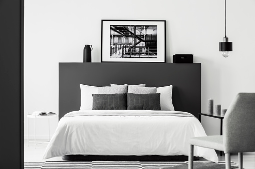 Poster on black bedhead of white bed in contrast bedroom interior with grey chair and lamp