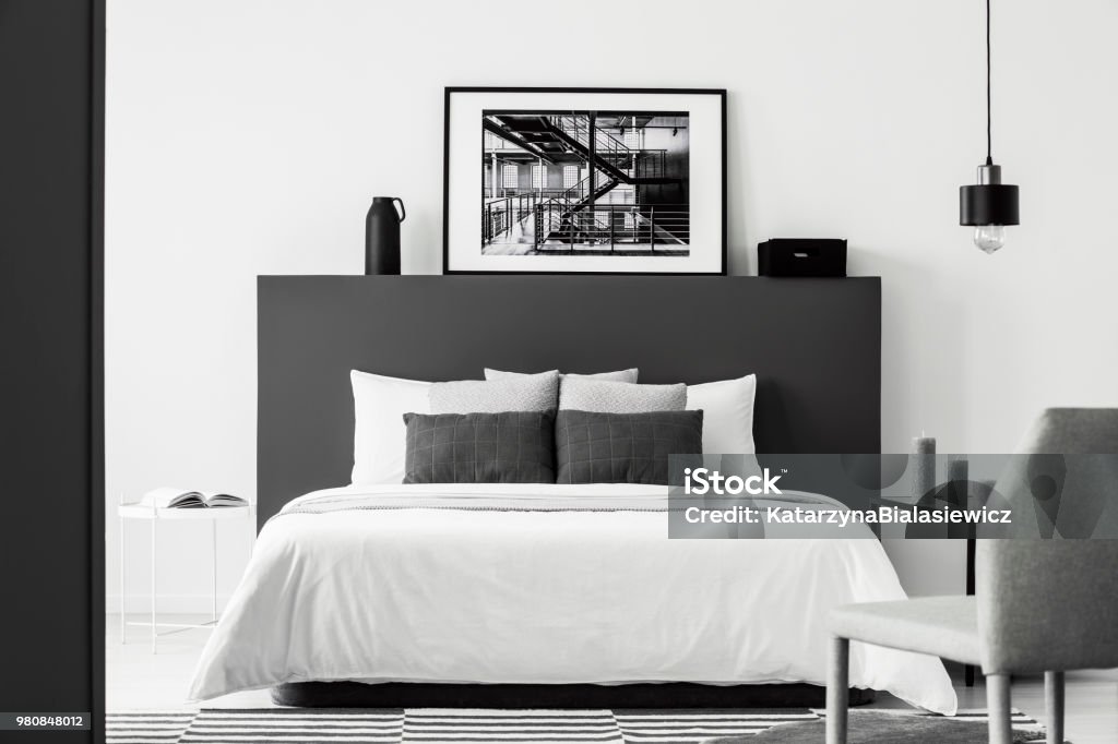Poster in contrast bedroom interior Poster on black bedhead of white bed in contrast bedroom interior with grey chair and lamp Black And White Stock Photo