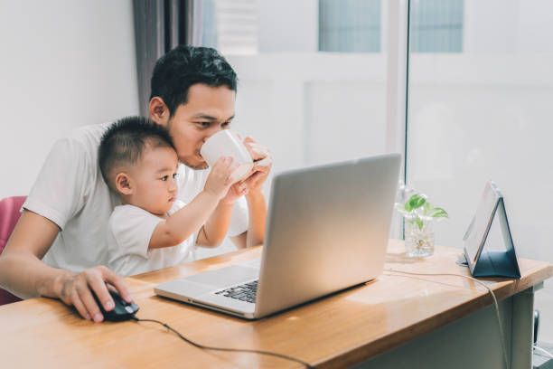 アジアの小さな息子は、幸せな家庭を職場でラップトップ コンピューターのノートブックで一緒に在宅勤務概念間コーヒーを飲む父を助けます。 - family room ストックフォトと画像
