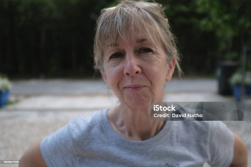 60 year old cute petite woman outdoors horizontal causal portrait Blode hair with bangs in late afternoon gravel driveway with forrest looking background. Health active lifestyle. Environmental portrait. Retirement and financial planning. Women Stock Photo