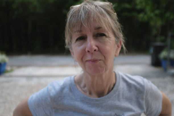 60 year old cute petite woman outdoors horizontal causal portrait Blode hair with bangs in late afternoon gravel driveway with forrest looking background. Health active lifestyle. Environmental portrait. Retirement and financial planning. 55 59 years stock pictures, royalty-free photos & images