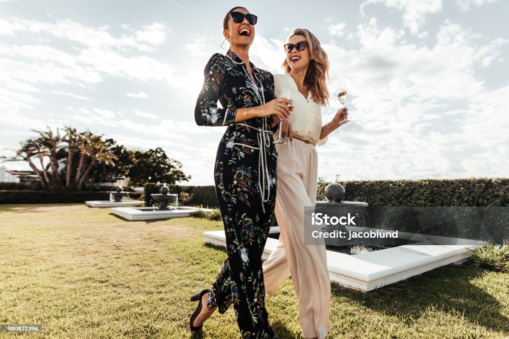 Stylish female friends walking together Portrait of two female friends walking on the lawn and laughing. Young women with wine walking together outside and smiling. Women Stock Photo