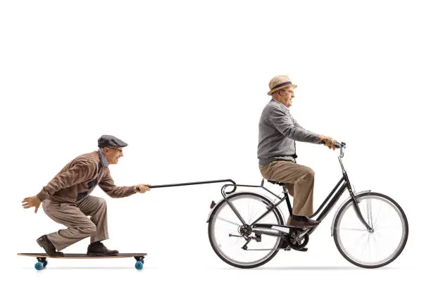 Photo of Senior riding a bike with another senior riding a longboard and pulling himself with a cane