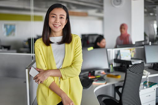 Portrait of confident young businesswoman at work place. Smiling female professional is holding mobile phone at desk. She is wearing smart casuals in office.