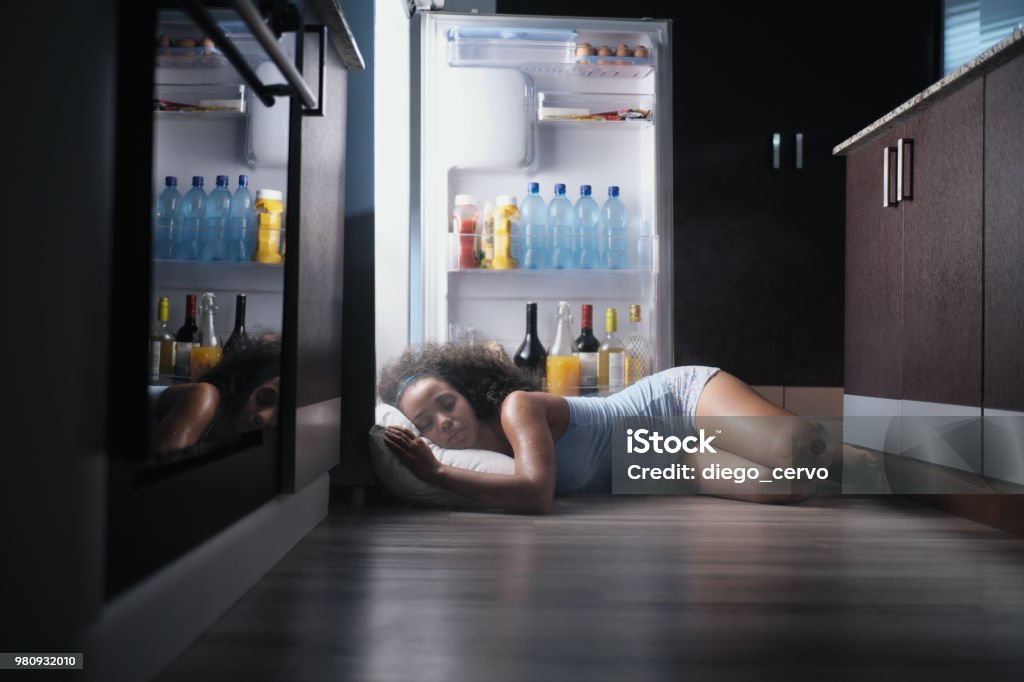 Black Woman Awake For Heat Wave Sleeping in Fridge Young hispanic woman suffering for summer heat and lack of air conditioning at home. Black girl covered with sweat sleeping on floor with head inside fridge. Heat - Temperature Stock Photo