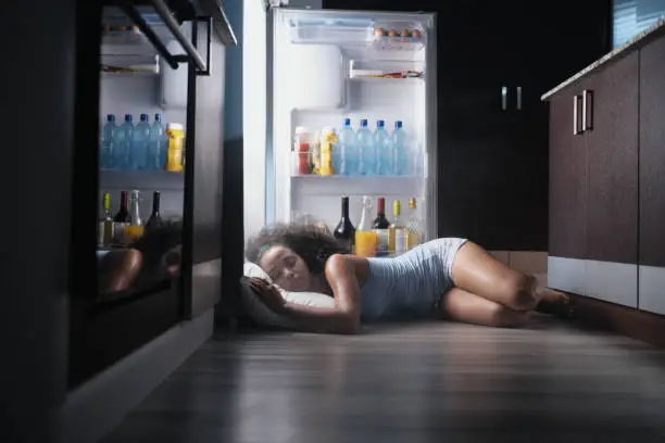 Young hispanic woman suffering for summer heat and lack of air conditioning at home. Black girl covered with sweat sleeping on floor with head inside fridge.