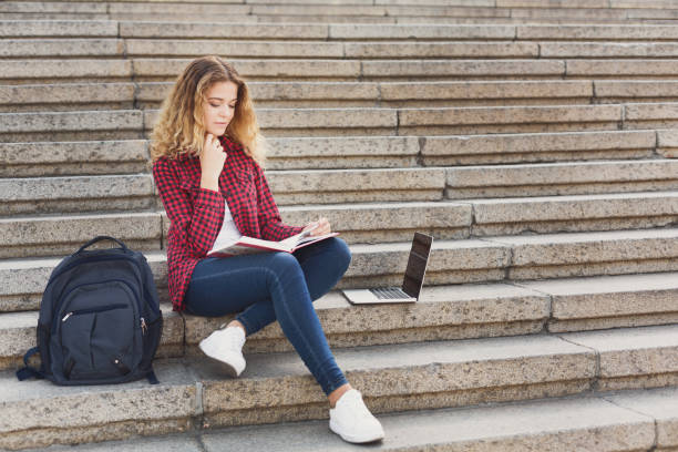 istock Smiling female student sitting on the stairs using laptop outdoo 980954374