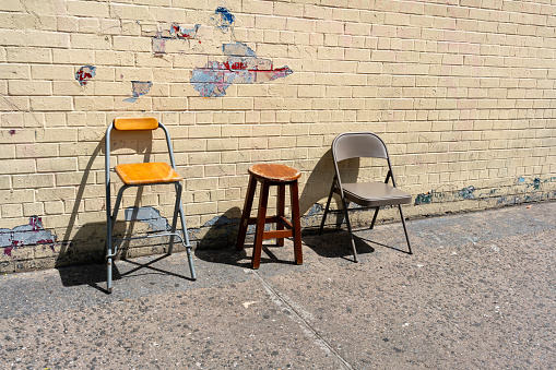 A picture of two chair and a stool by a wall on a sidewalk.
