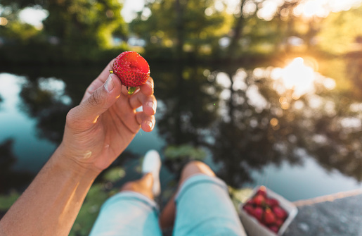 Personal perspective of a young man eating strawberries by river in Sweden during sunset. He is holding up a berry with a missing bite.
