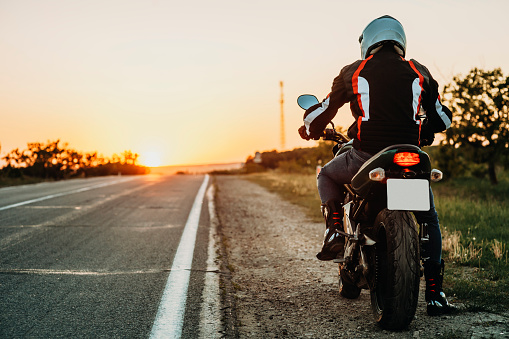 Back view of male in protective jacket, boots and helmet riding off roadside at sunset on empty highway backlit background