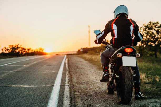 Man on motorbike riding on roadside Back view of male in protective jacket, boots and helmet riding off roadside at sunset on empty highway backlit background biker photos stock pictures, royalty-free photos & images