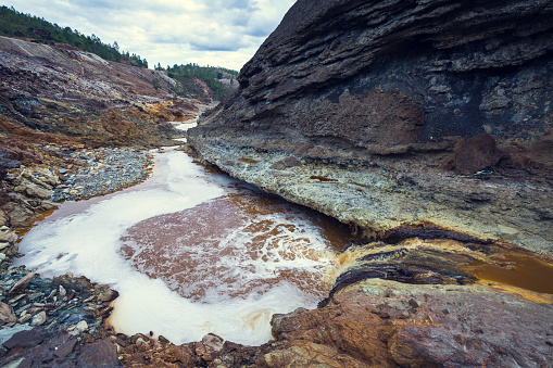 Stream of acid orange water with low PH between the cliff with black and red rocks, with the pine forest in the background in the old Zaranda mine, Spain