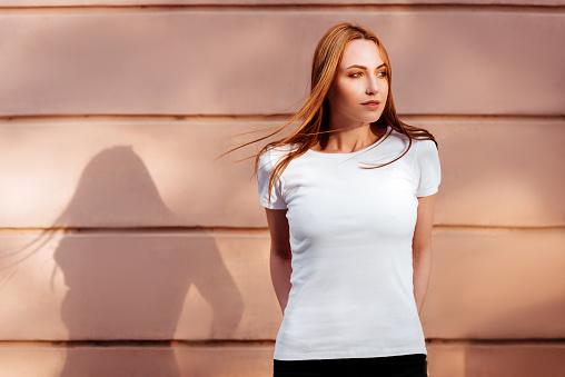 Girl wearing blank white t-shirt, jeans posing against rough street wall, minimalist urban clothing style.