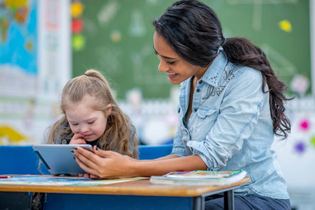 Teaching elementary school girl with a tablet A young teacher shows her student something on a tablet at her classroom desk. Her student is elementary aged and has Down syndrome. She shows interest in the tablet. assistant stock pictures, royalty-free photos & images
