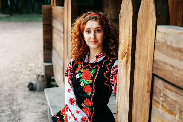 Young Slavonic woman in traditional embroidered costume and red shoes sitting on the porch