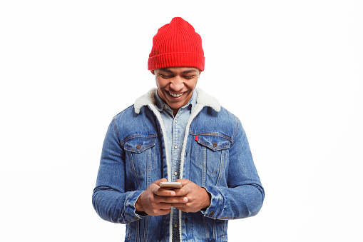 Cheerful black male model in denim jacket surfing new smartphone and looking happy isolated on white.