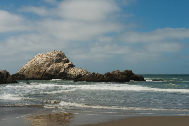 This is a color photograph of the Ocean Beach landscape in San Francisco, California during the summer. Rugged rocks stick out of the water along the shoreline of the Pacific ocean.