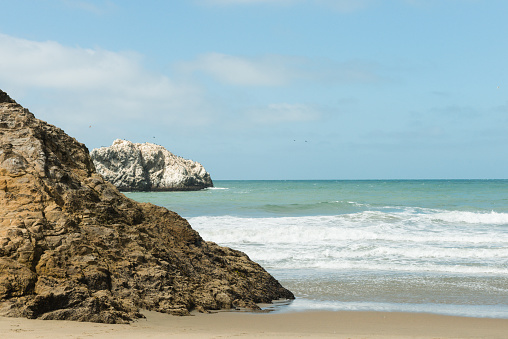 This is a color photograph of the Ocean Beach landscape in San Francisco, California during the summer. Rugged rocks stick out of the water along the shoreline of the Pacific ocean.