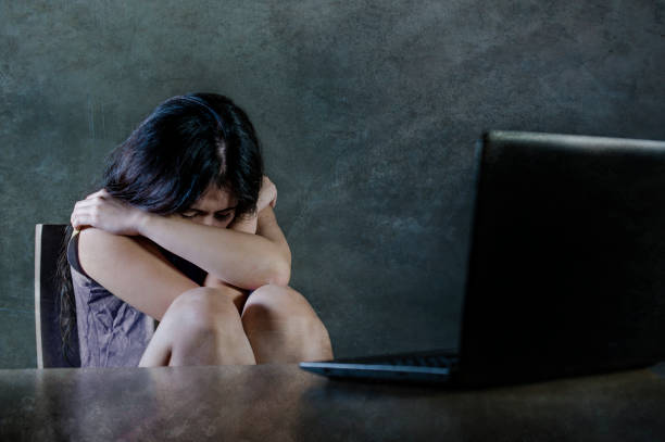 dramatic portrait of young sad and scared woman holding head stressed and worried looking at laptop computer isolated on dark background in cyber bullying and internet problem stock photo