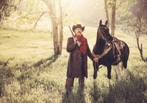 Cowboy holds up a revolver in one hand standing in front of the white background