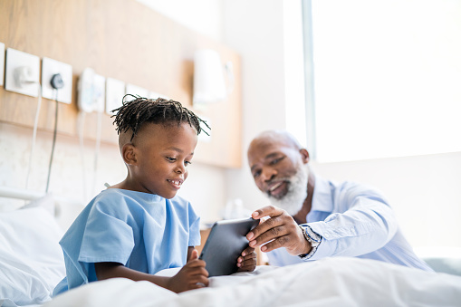 Grandfather showing digital tablet to sick grandson. Smiling male patient is using wireless computer on bed. They are at hospital ward.