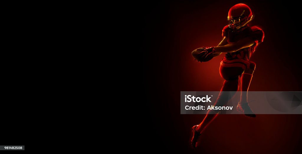 American football player with ball on a dark red background American football player with ball on a dark red background with red glow. Sportsman dressed in american football sport uniform Running Stock Photo