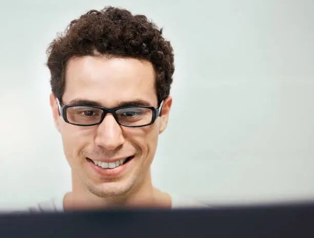 A good-looking young man of Spanish or Portuguese descent smiles at his pc monitor.