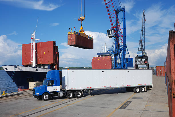 A large industrial crane lifts a red cargo platform with a worker standing on it at a shipping port. Stacked colorful freight containers and a barbed-wire fence are visible