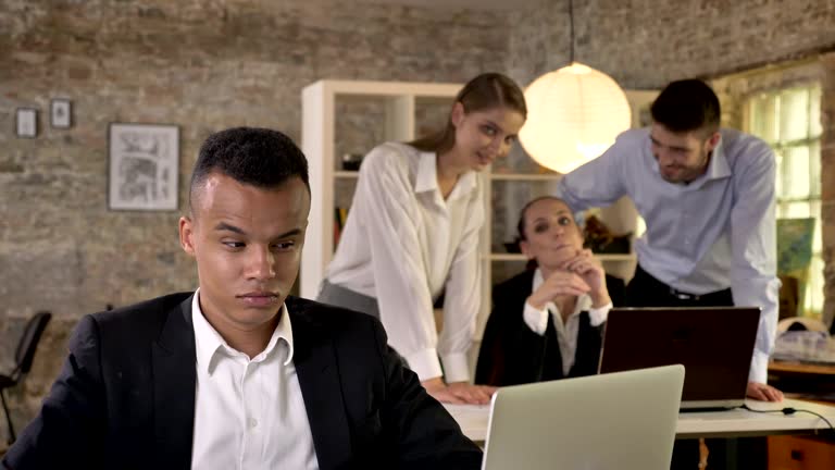 Young afro american businessman is listening how his colleagues on background gossip about him, racism concept, bulling concept