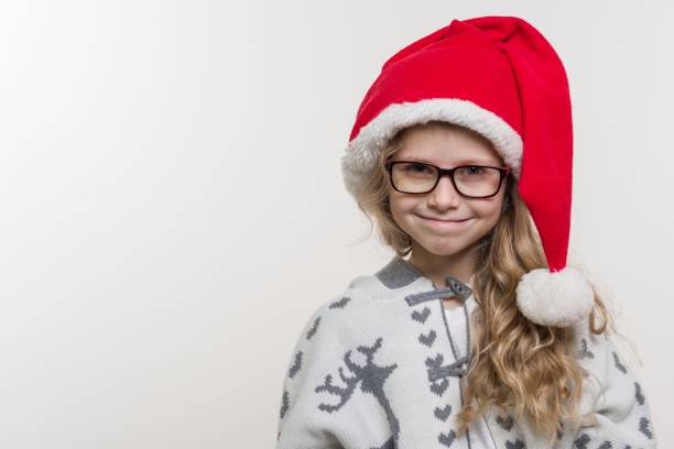 vacaciones de invierno - un retrato abierto de divertida niña de 7 años en santa claus sombrero, gafas, suéter tejido con patrón tradicional - ciervo, sobre fondo claro. copia espacio - eyewear child glasses 6 7 years fotografías e imágenes de stock