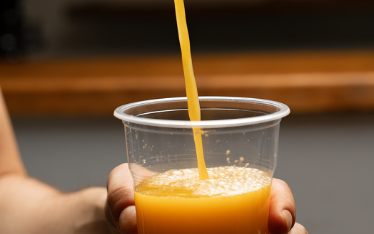 Freshly squeezed orange juice stream pouring into take-out plastic disposable glass held in a hand of a young man, close-up shoot with selective focus. Contains bokeh in the background for copy space.