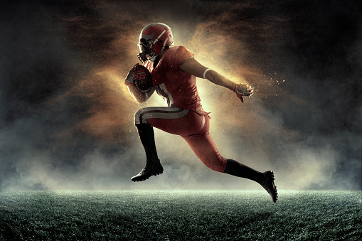 American football player jumps and catches the ball in flight in professional sport stadium with smoke and fog. Sportsman dressed in red sport uniform