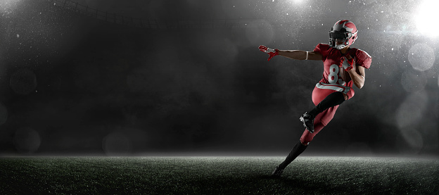American football player jumps and catches the ball in flight in professional sport stadium with fog. Sportsman dressed in red sport uniform