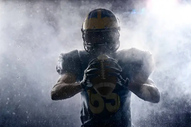 American football player in a haze and rain on black background. Portrait. Athlete dissecting white smoke and water drops. Sportsman shines in the rays of light