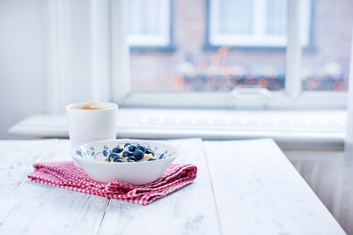 Plate with muesli and berries, coffee in a cup for breakfast. Napkin in a red cage on a white wooden table by the window. Free space for text.