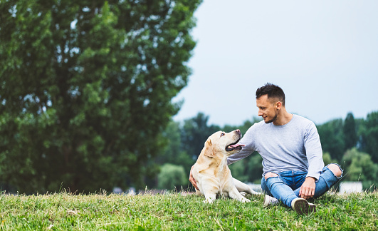 Urban young man is sitting on grass and hugging with his Labrador Retriever in nature.